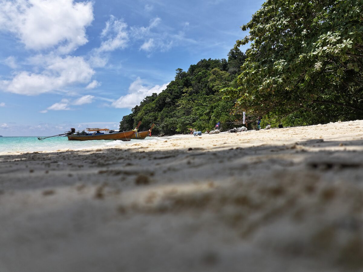 Low view across a white beach: longtail boats on turquoise water, jungle-covered hills behind.
